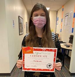 A woman wearing a mask is holding a certificate in a hallway.