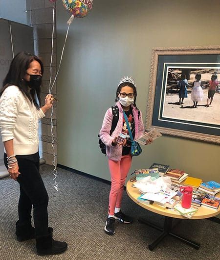 A woman and a girl wearing masks are standing next to a table holding balloons.