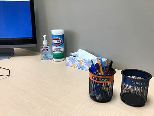 A desk with a computer , a pen holder , a bottle of hand sanitizer , and a tissue box.