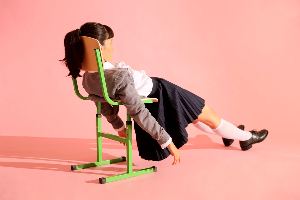 A girl in a school uniform is sitting on a green chair on a pink background.