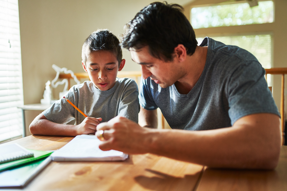 A man is helping a young boy with his homework at a table.