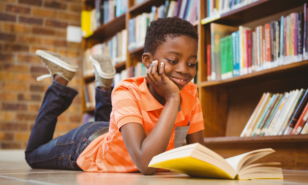 A young boy is laying on the floor reading a book in a library.