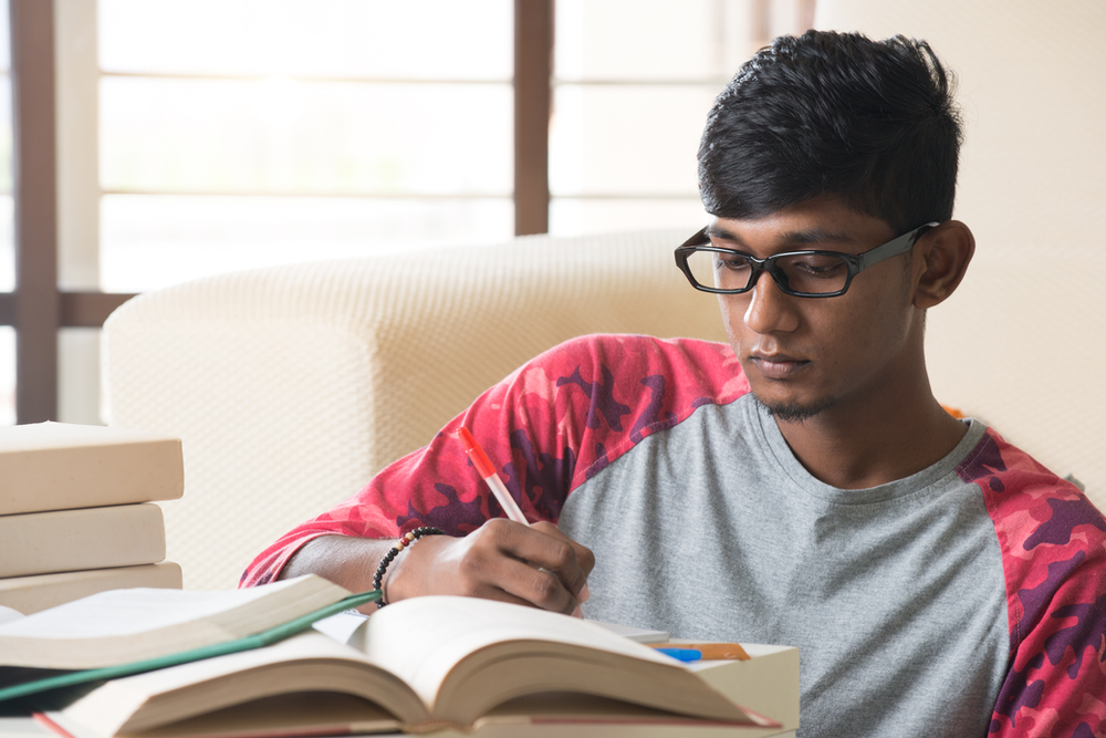 A young man is sitting at a table reading a book and writing in a notebook.