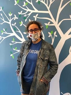 A woman wearing a mask and a denim jacket is standing in front of a tree.
