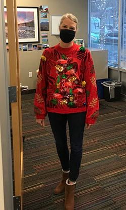 A woman wearing a christmas sweater and a mask is standing in a room.