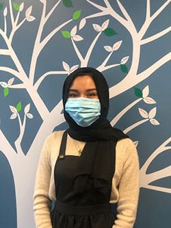 A woman wearing a face mask is standing in front of a tree.