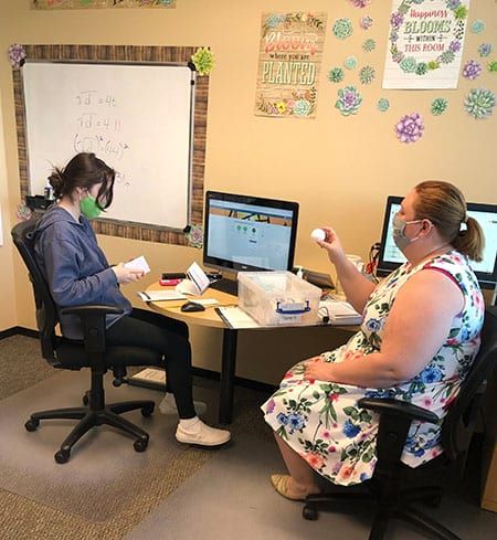 Two women wearing masks are sitting at a desk with computers.