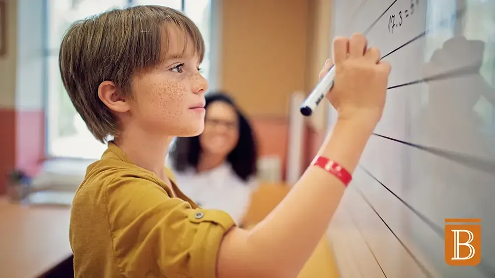 boy working at whiteboard with teacher in the distance