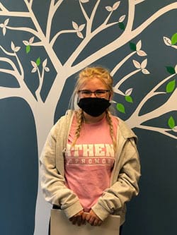 A girl wearing a mask and a pink shirt is standing in front of a tree.
