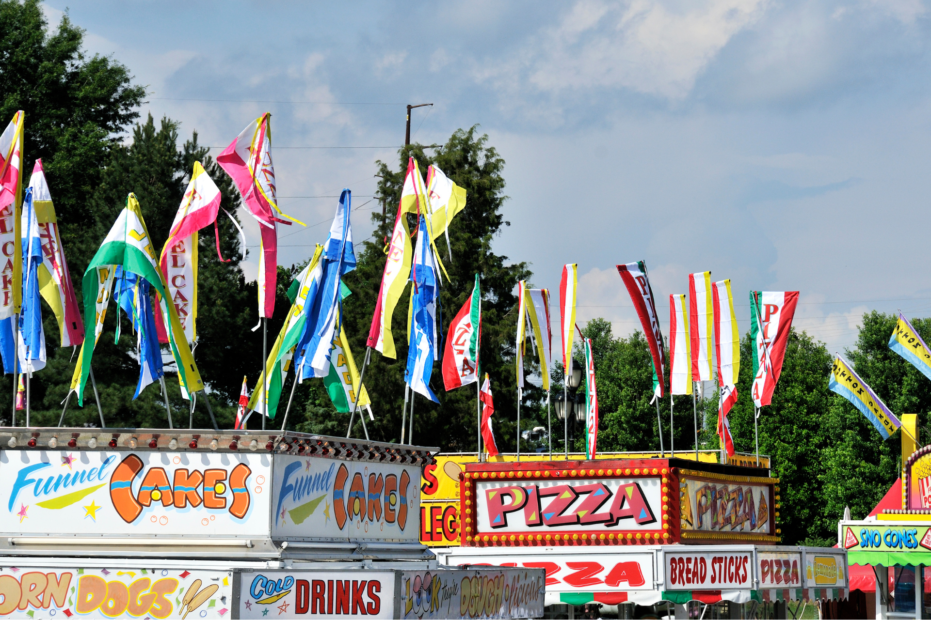Food vendor booths at an outdoor fair, flags overhead, signs for funnel cakes, pizza, and drinks.