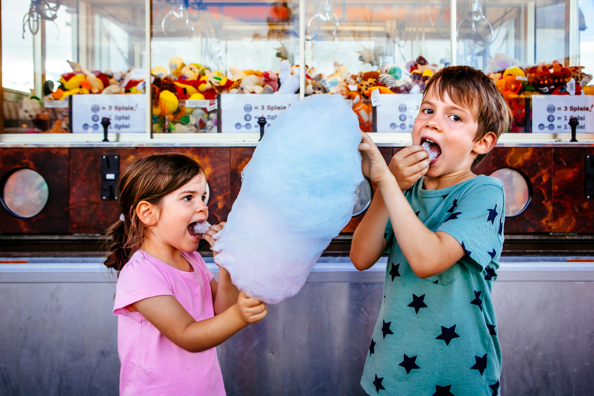 Two children eating a large, blue cotton candy outside a prize booth.
