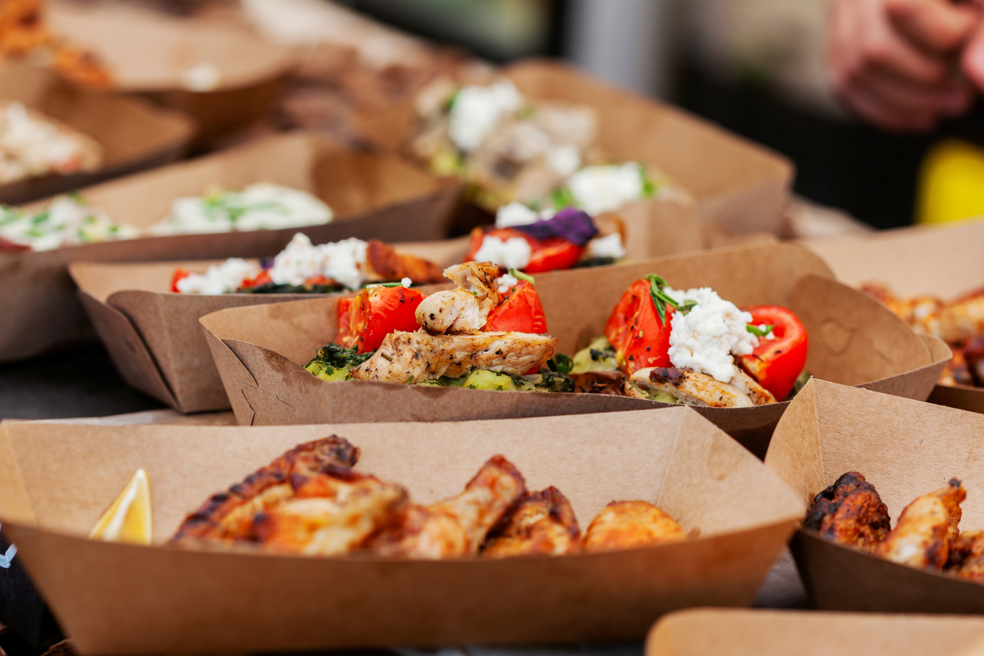 Food in brown paper trays, including chicken, tomatoes, and other ingredients, at a market.
