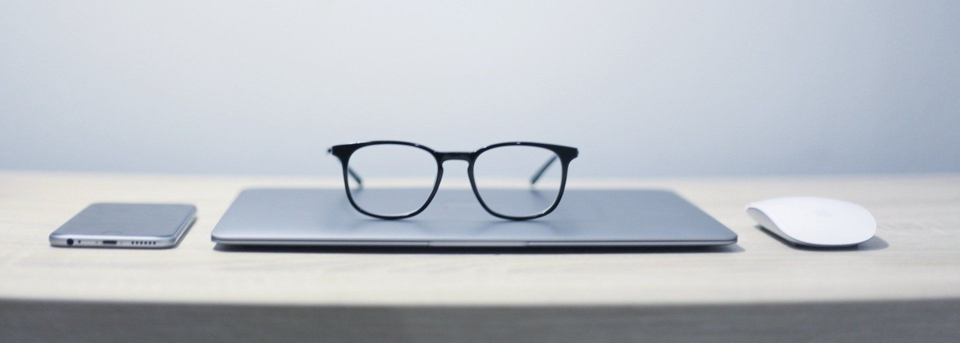 Een bril met zwart montuur rust op een dichtgeklapte laptop, met aan weerszijden een telefoon en een witte muis.