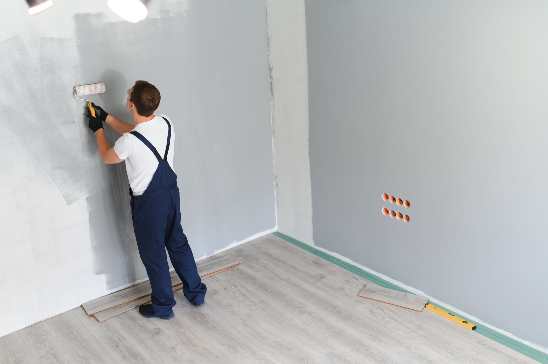 Person in blue overalls painting a gray wall with a roller in a room with gray flooring.