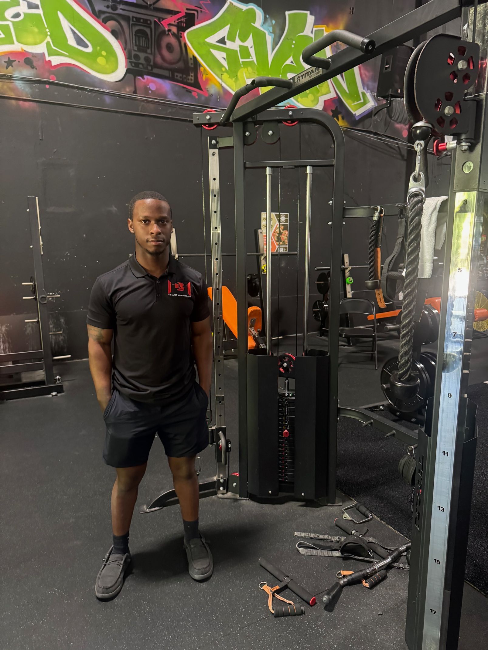Man in gym standing next to cable machine. Black outfit, hands in pockets. Graffiti background.
