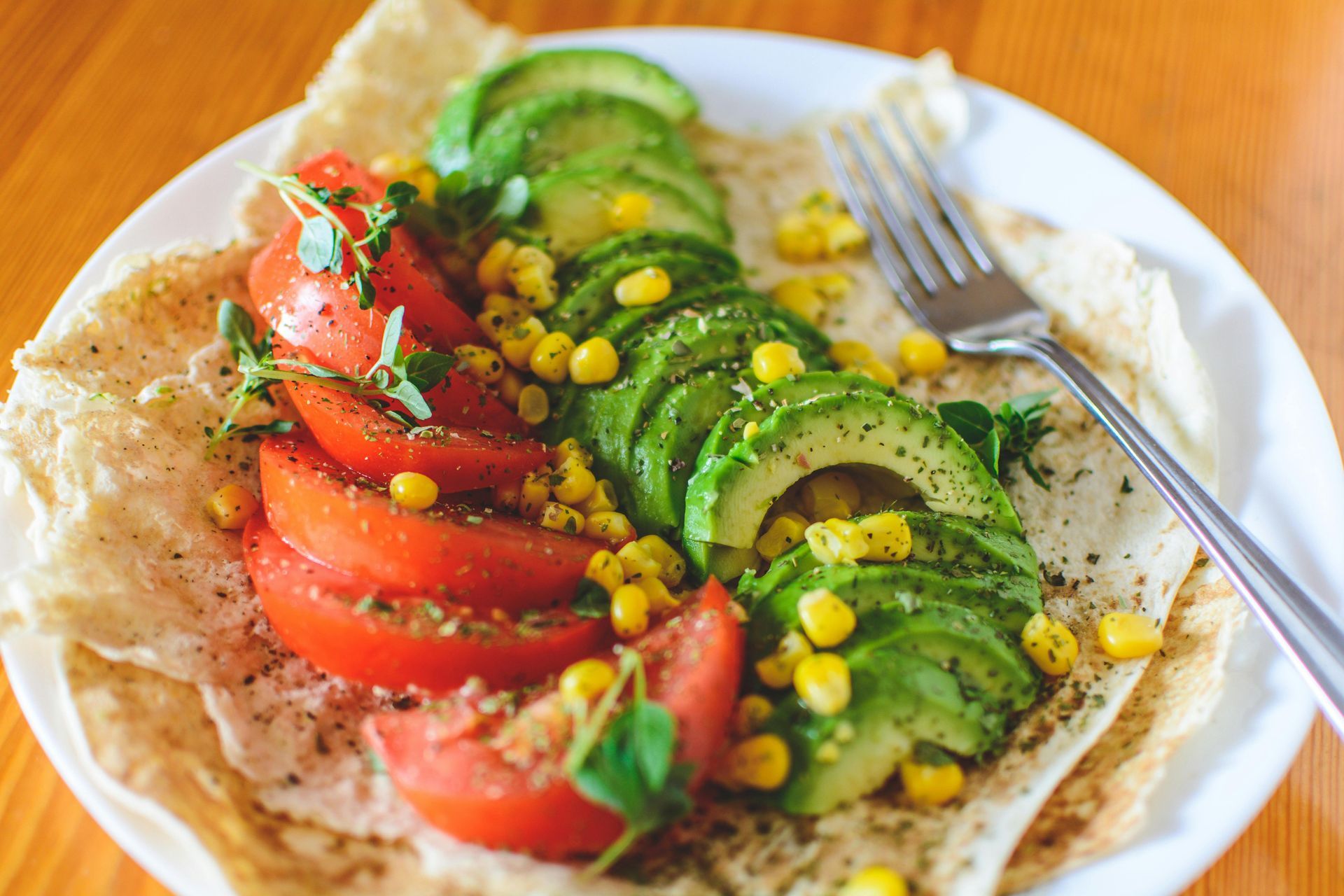 Plate of sliced avocado, tomatoes, and corn on a flatbread, with a fork.