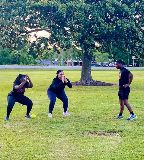 Three people exercising in a park: two doing squats, and one watching. Green grass, tree, and blue sky.