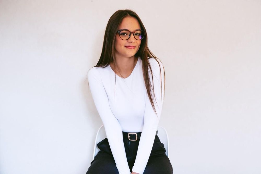 A person with long brown hair wearing glasses and a white long-sleeved shirt, sitting against a plain white background.