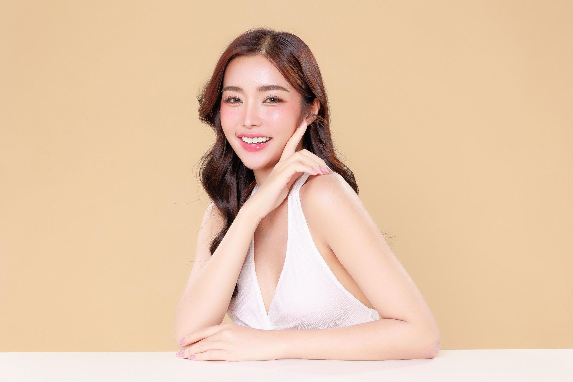 A woman in a white dress is sitting at a table with her hand on her face.