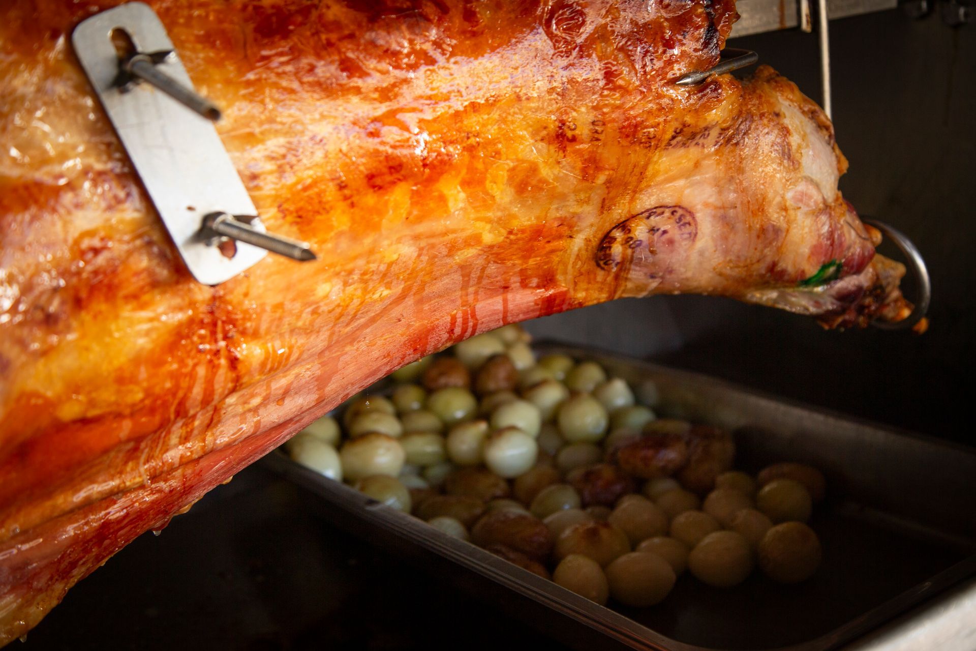 Roasted lamb on a rotisserie, with browned skin, over a pan of small onions.
