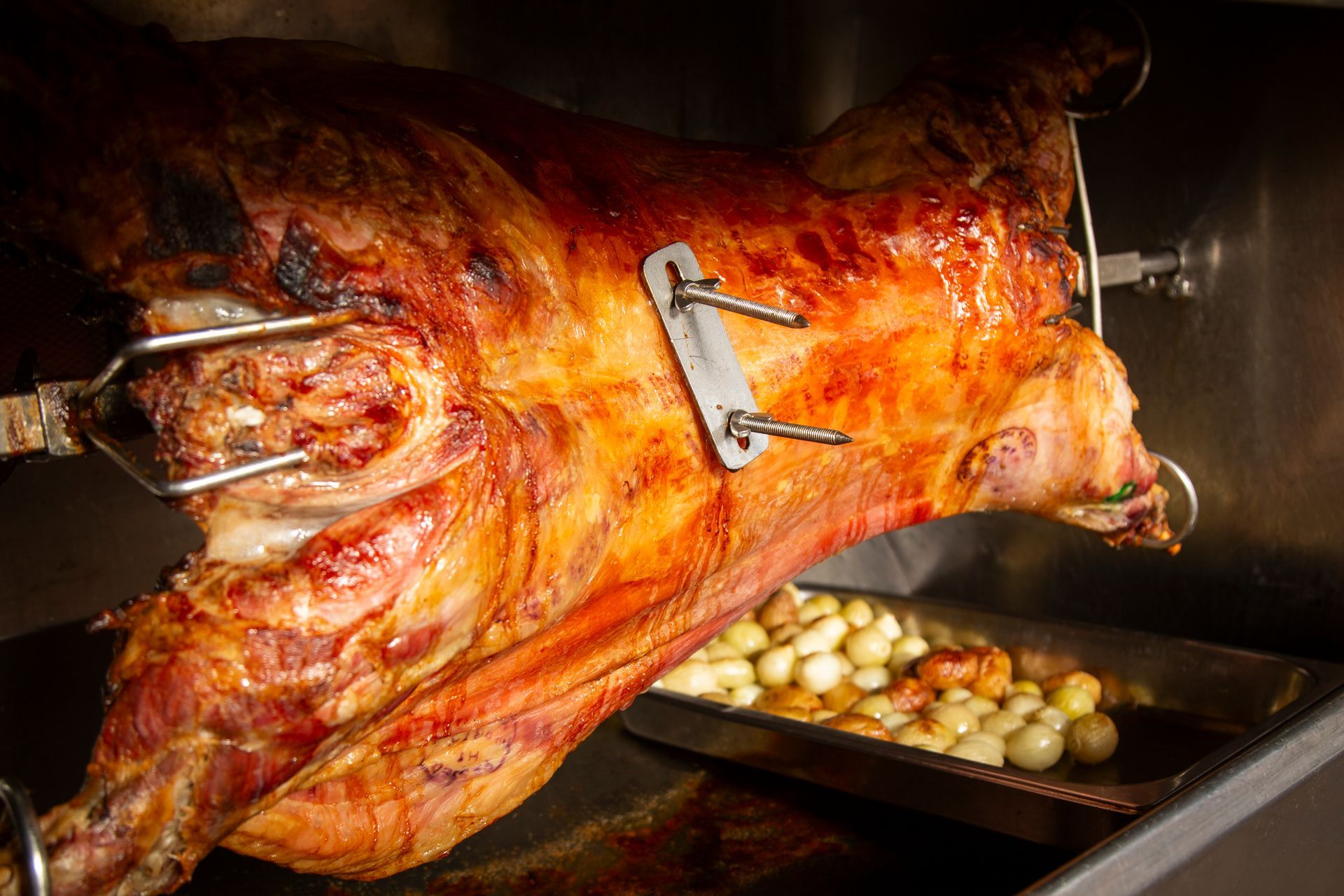 Whole roasted lamb turning on a spit, glistening with fat; roasting pan of onions below.