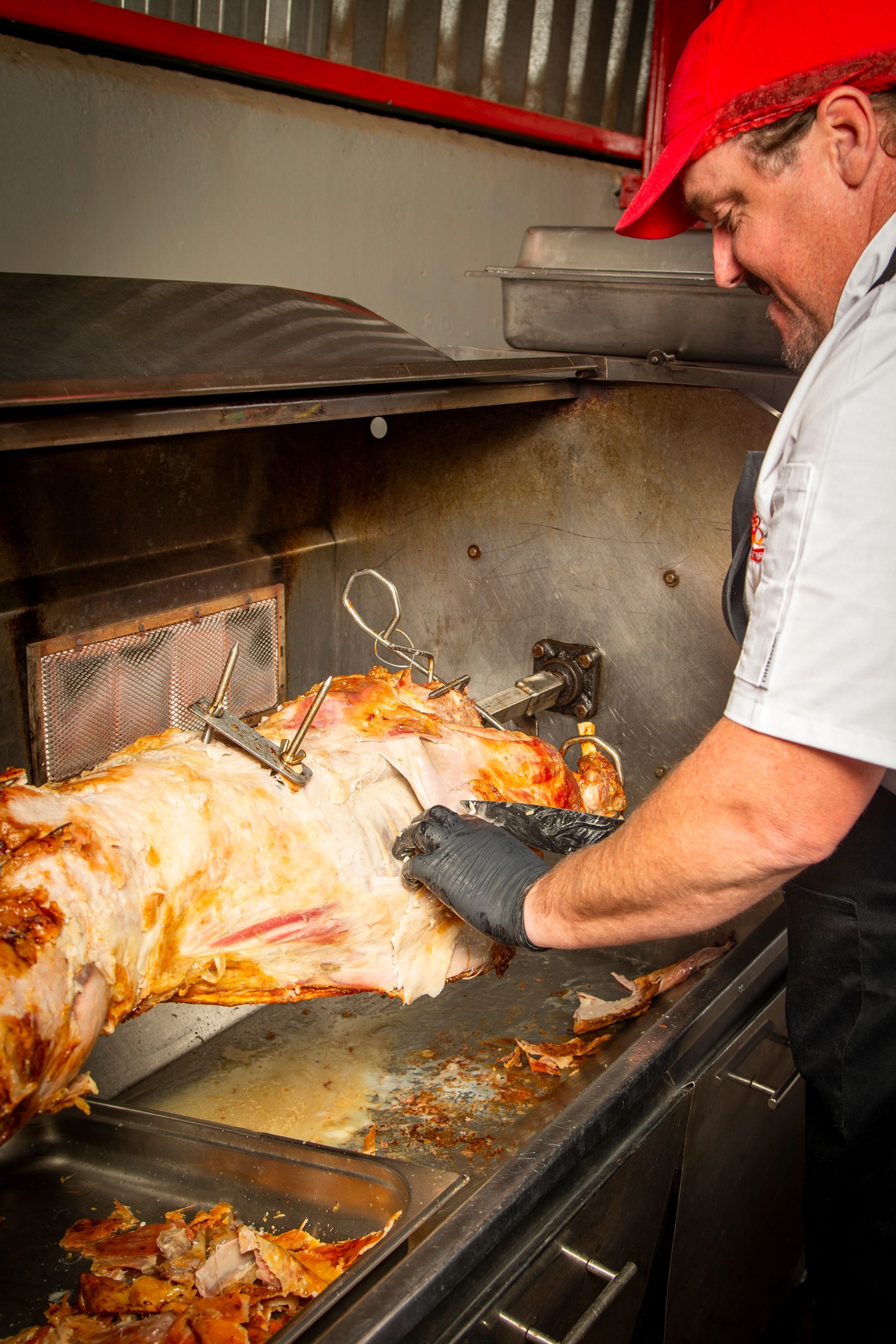 Man in red hat carving roasted meat from a spit.