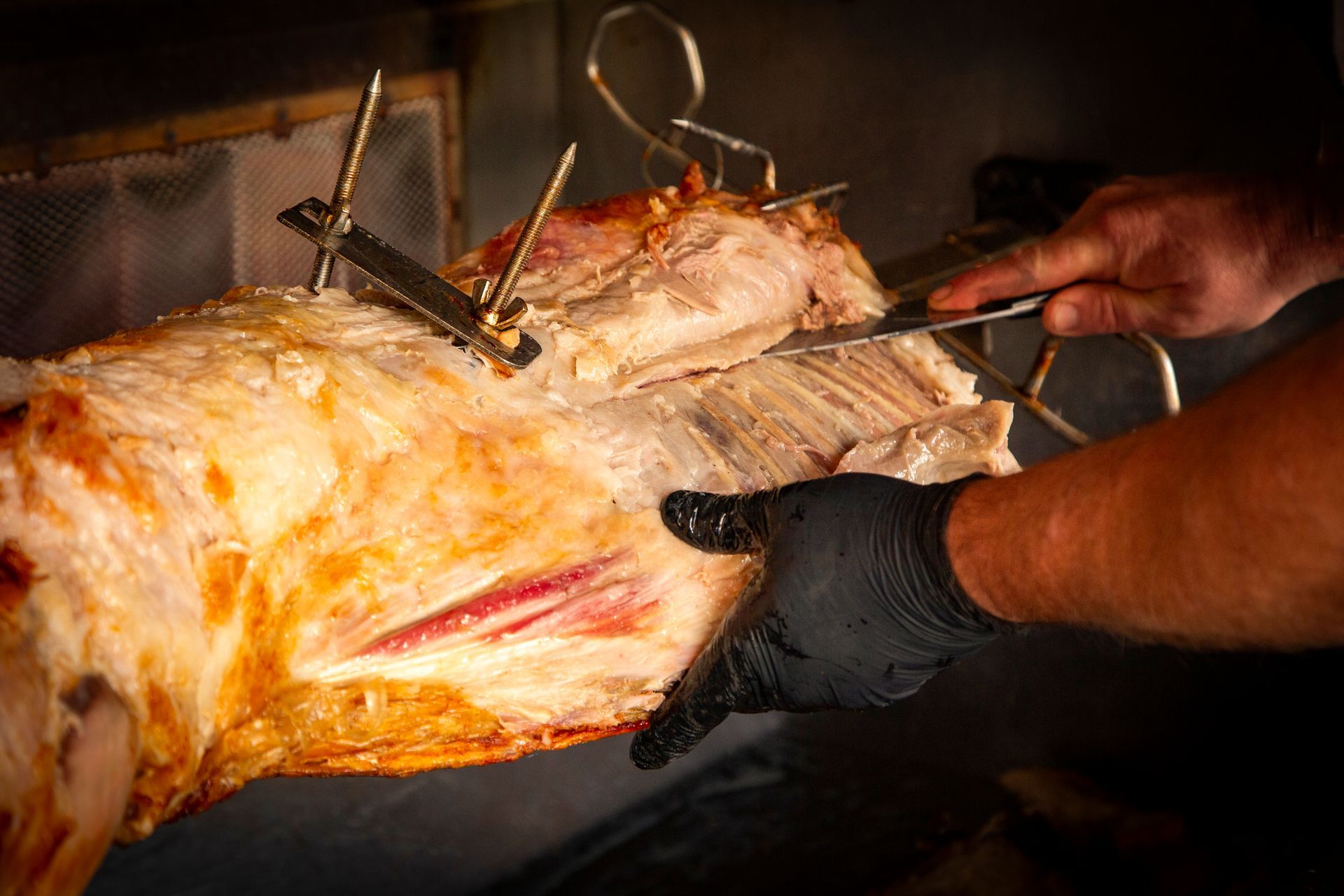 A person carving roasted meat on a rotating spit, wearing a black glove.