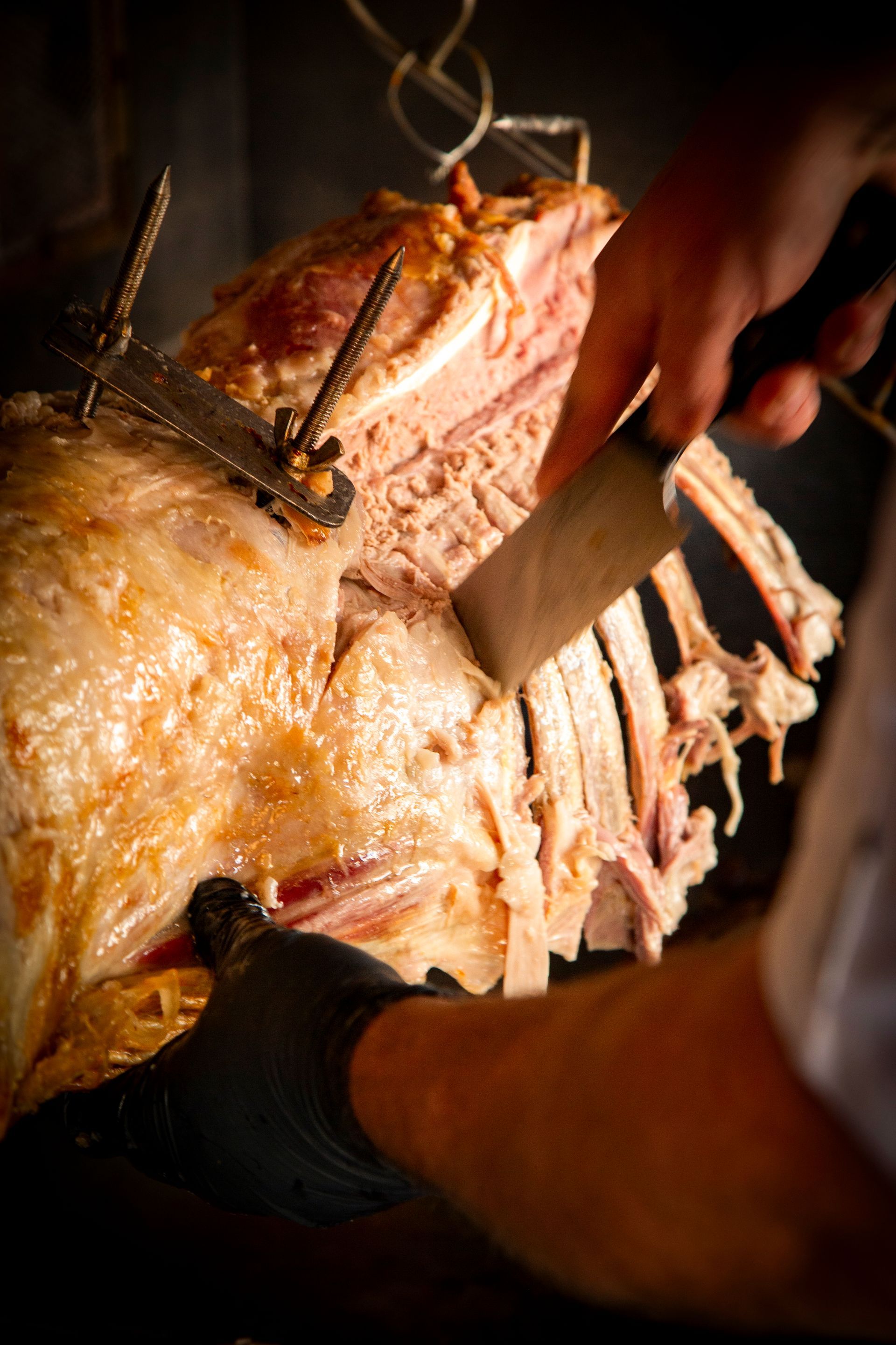 Butcher carving roasted meat, wearing a black glove, set against a dark background.