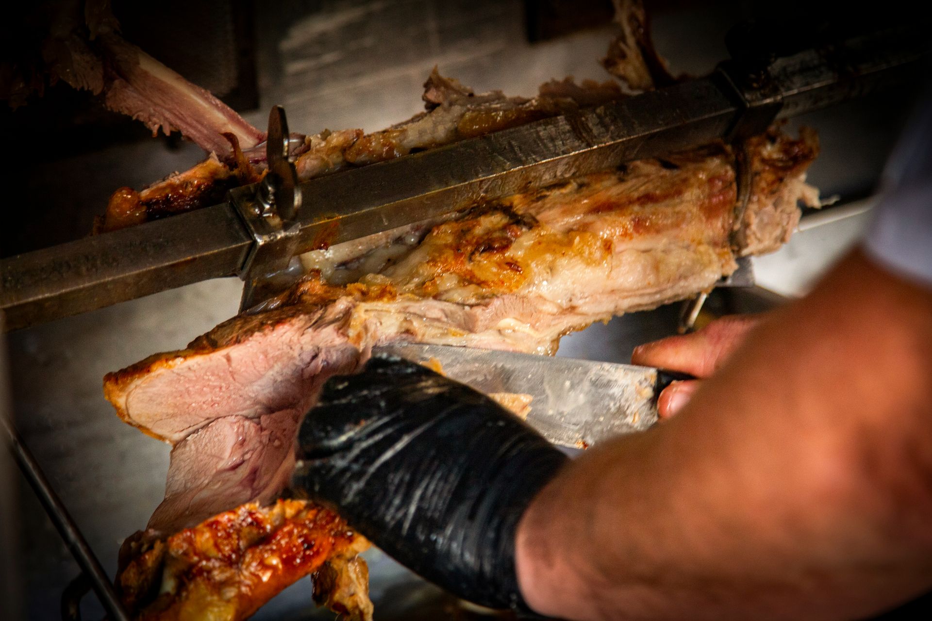 Person carving roasted meat from a spit, wearing a black glove, close-up.