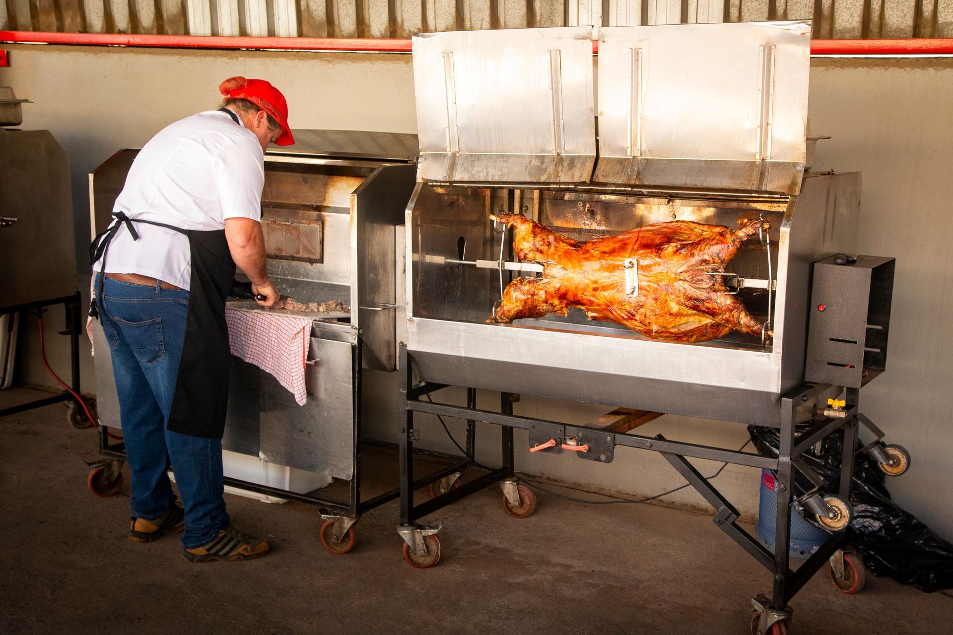 A man in a red hat tending to a roasting lamb on a rotisserie grill.