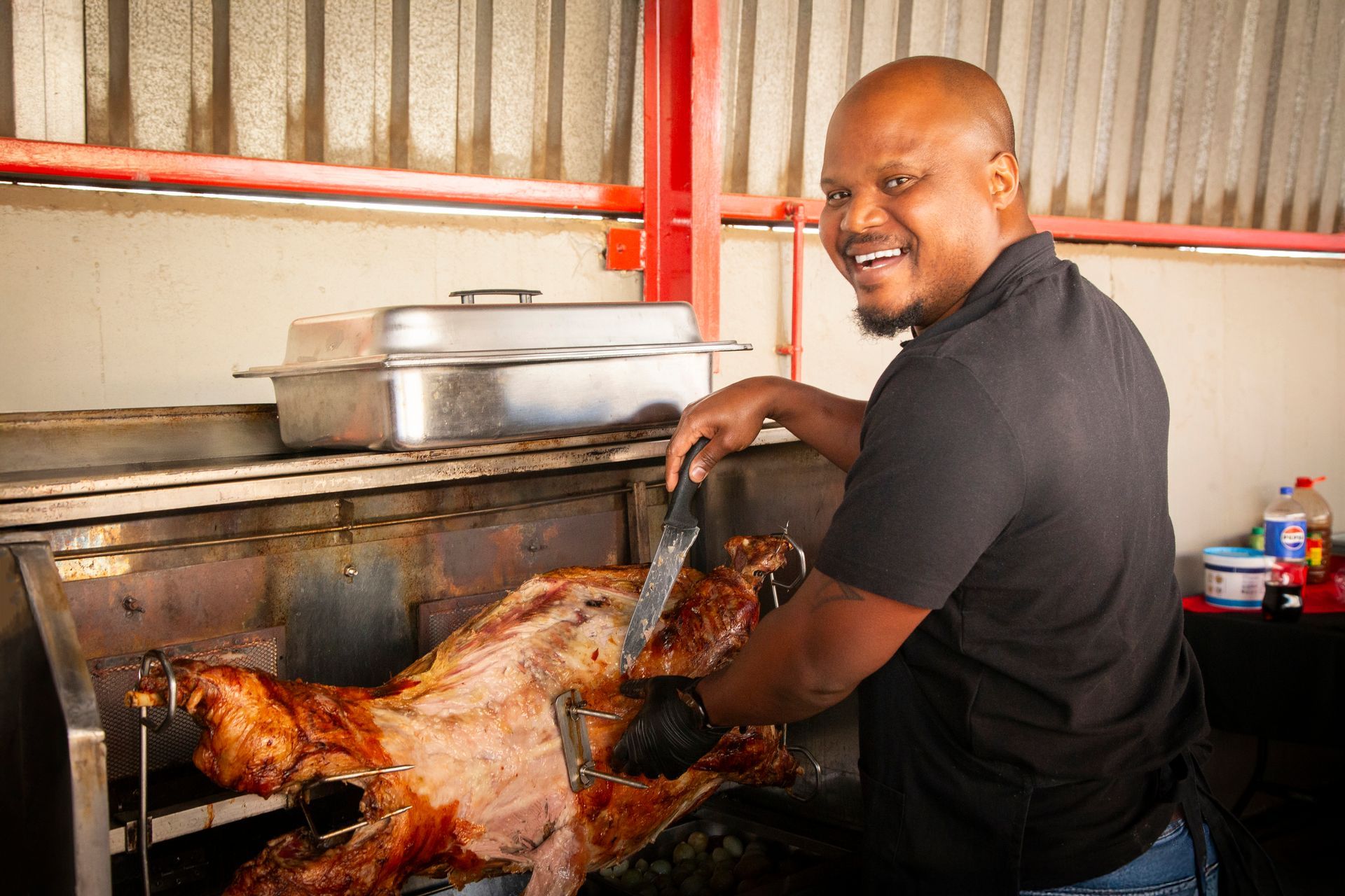 Man carving roasted meat, smiling. Outdoors with a red-piped ceiling, metal roasting spit.