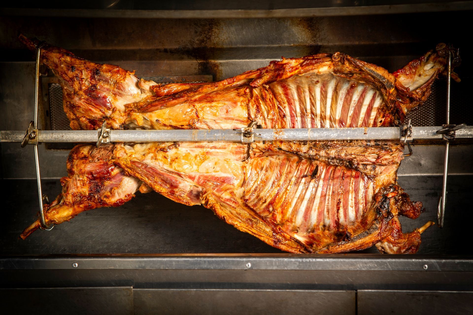 Roasted whole lamb on a rotisserie, golden-brown with visible rib bones, inside a metal oven.