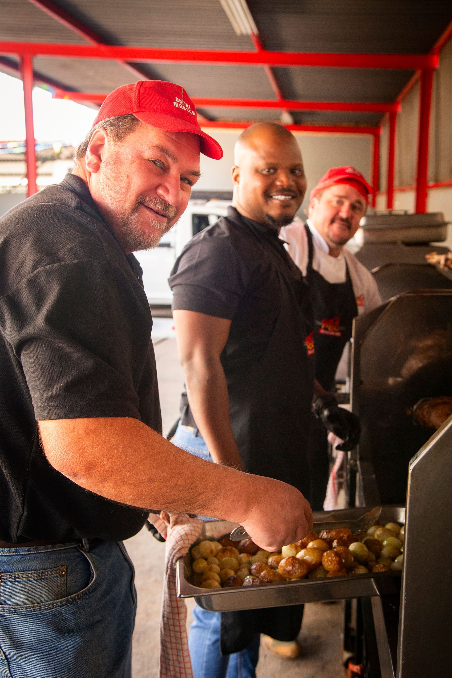 Three men smiling, cooking outdoors; one holds a tray of potatoes.