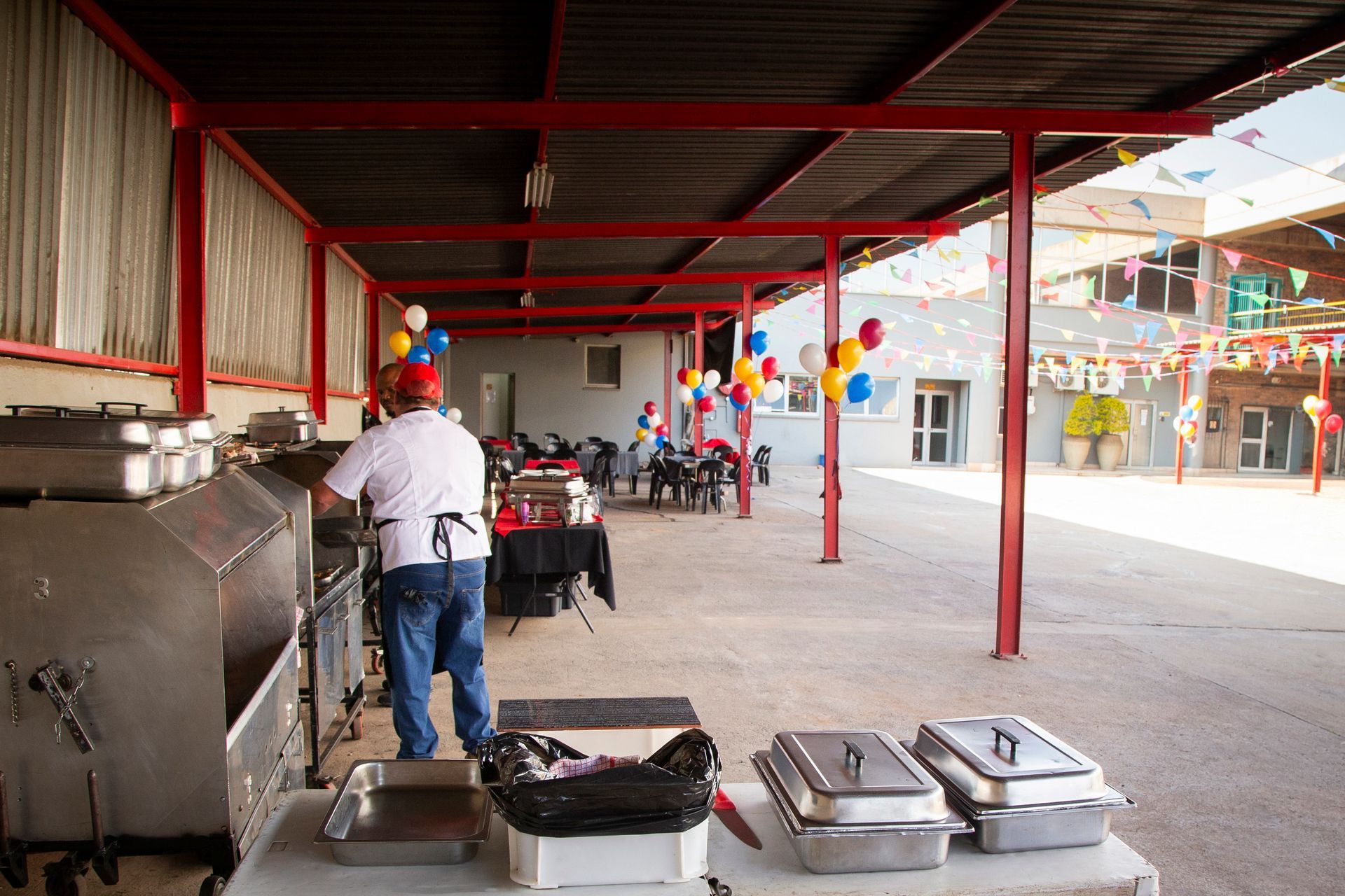 Man in apron preparing food at an outdoor event. Silver food warmers, red awning, balloons in background.