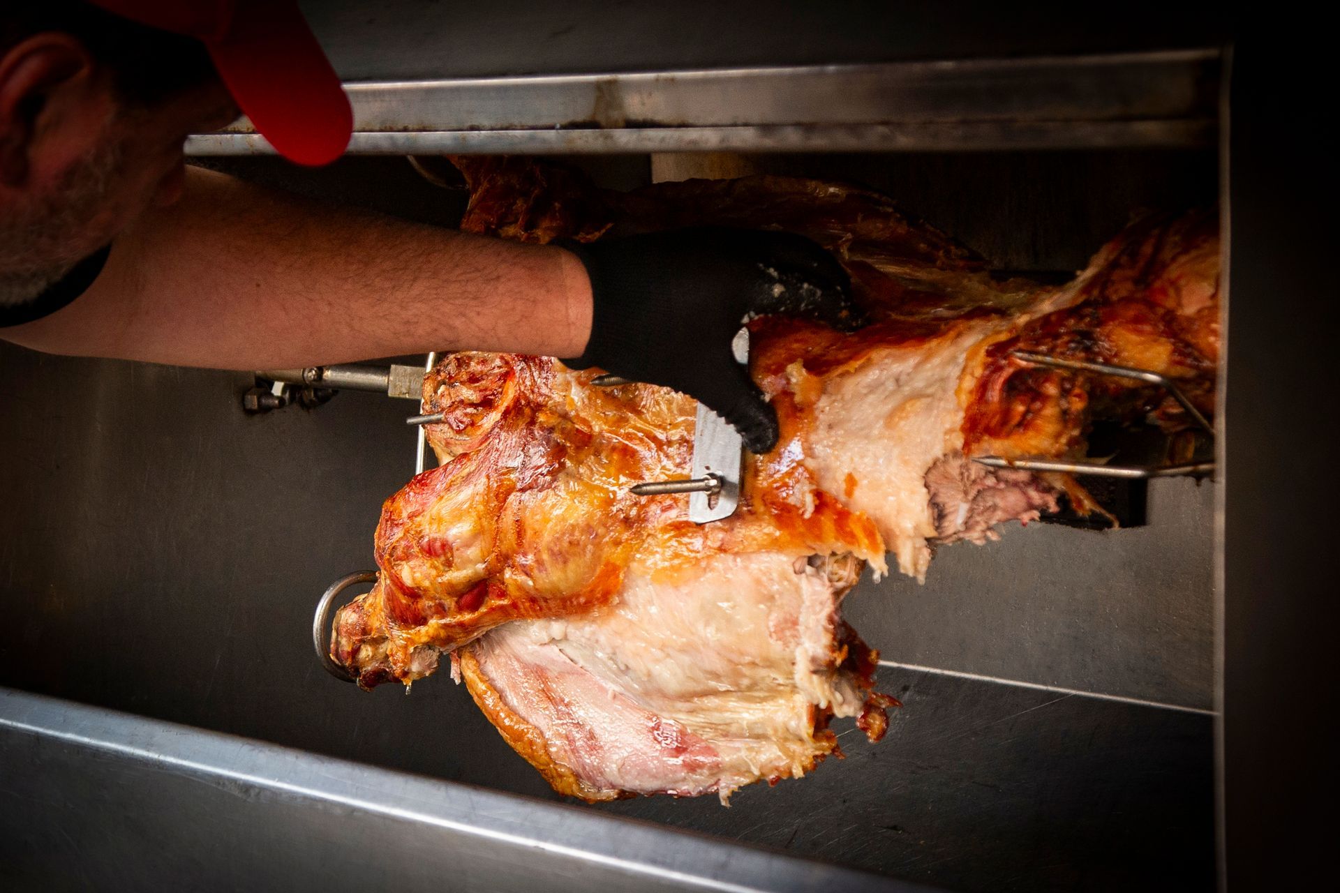 A person carves roasted meat on a rotisserie. The meat is golden brown with some raw flesh.
