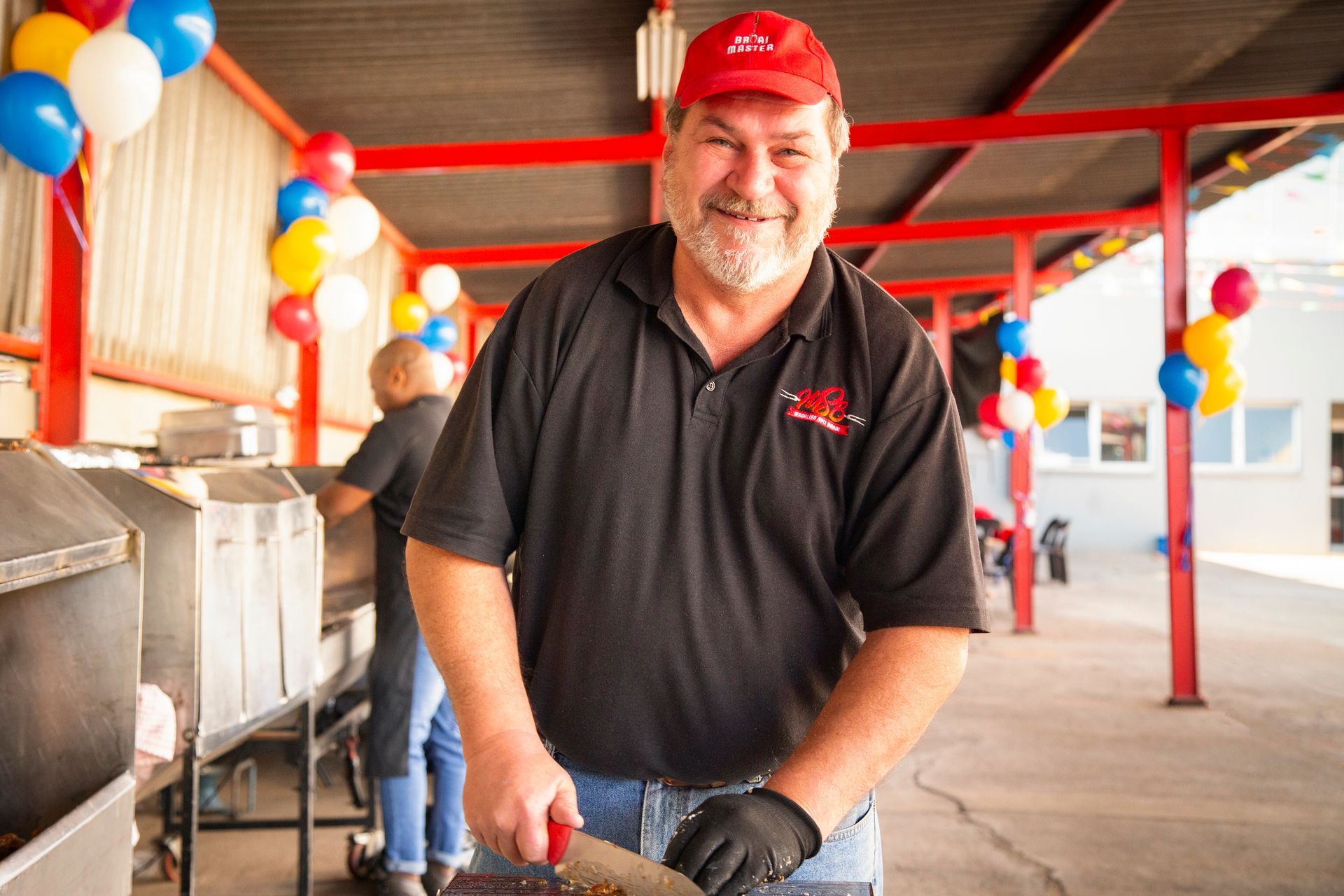 Man in red hat, smiling, carving meat at outdoor food station with balloons.