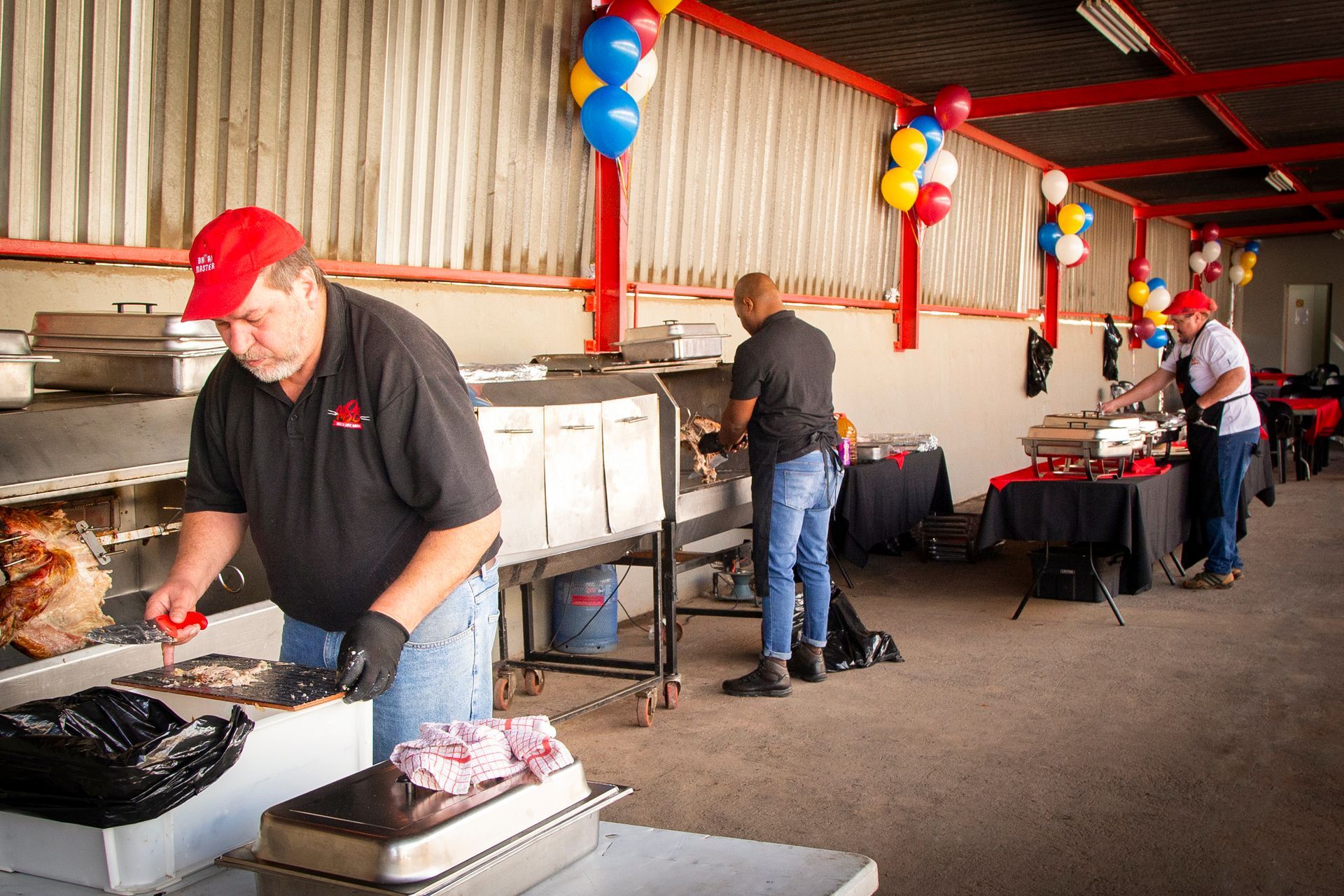 People preparing food at an outdoor event. Red and blue balloons decorate the open-air structure.