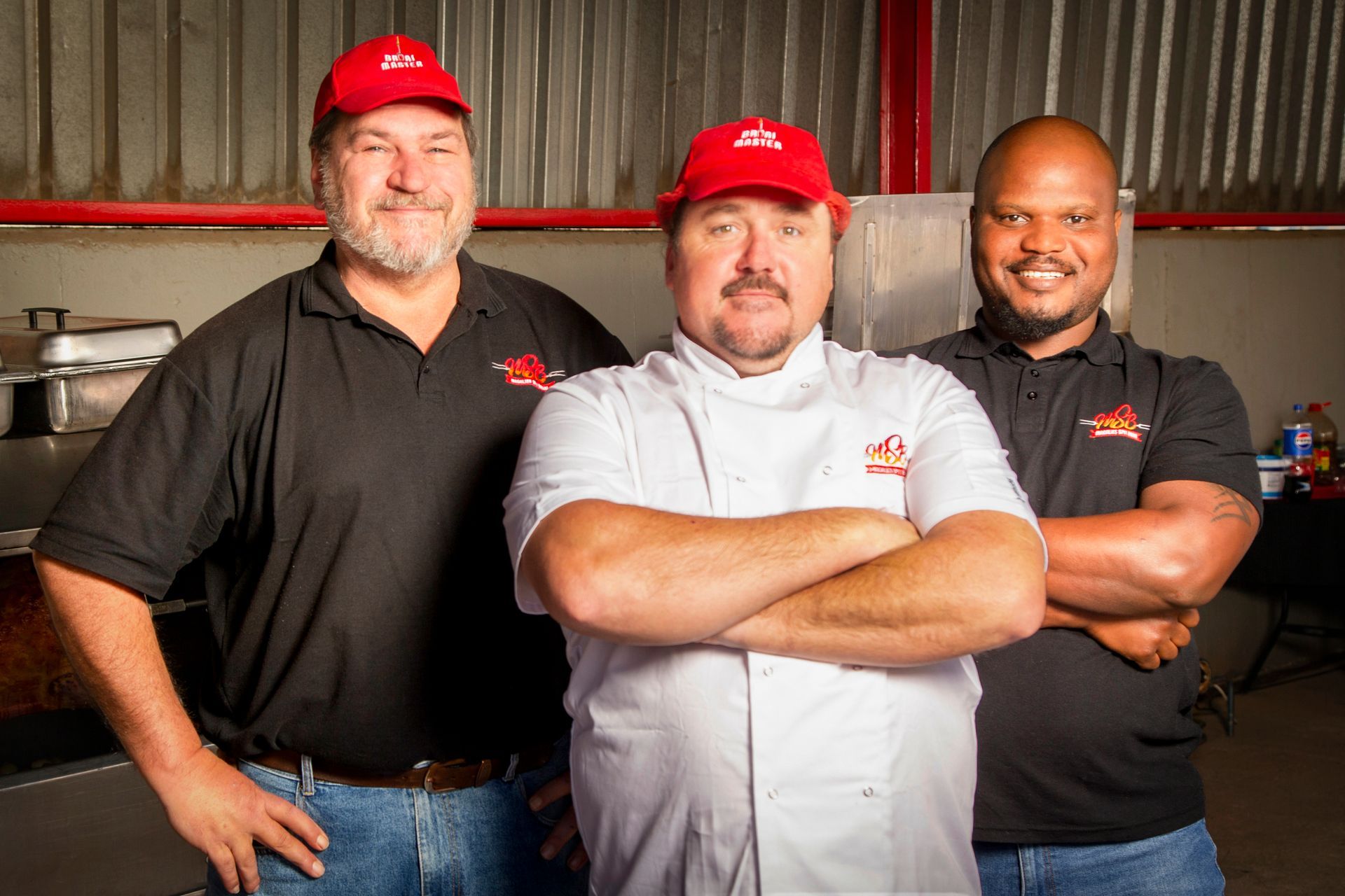 Three barbecue chefs wearing red hats and black shirts, one in a white chef's coat, smiling, posing indoors.