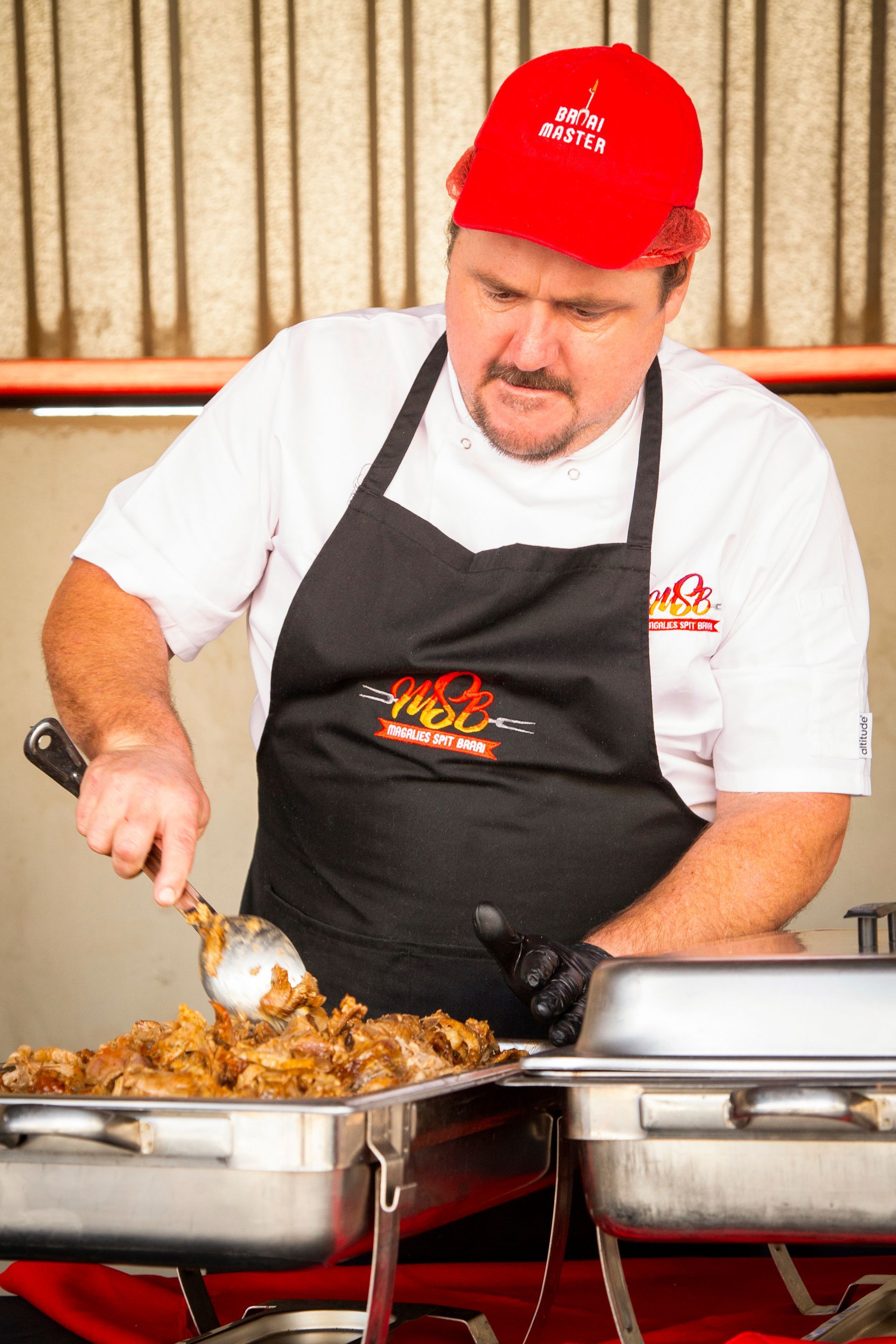 Chef in red cap, white shirt, black apron, scoops food from a metal tray.