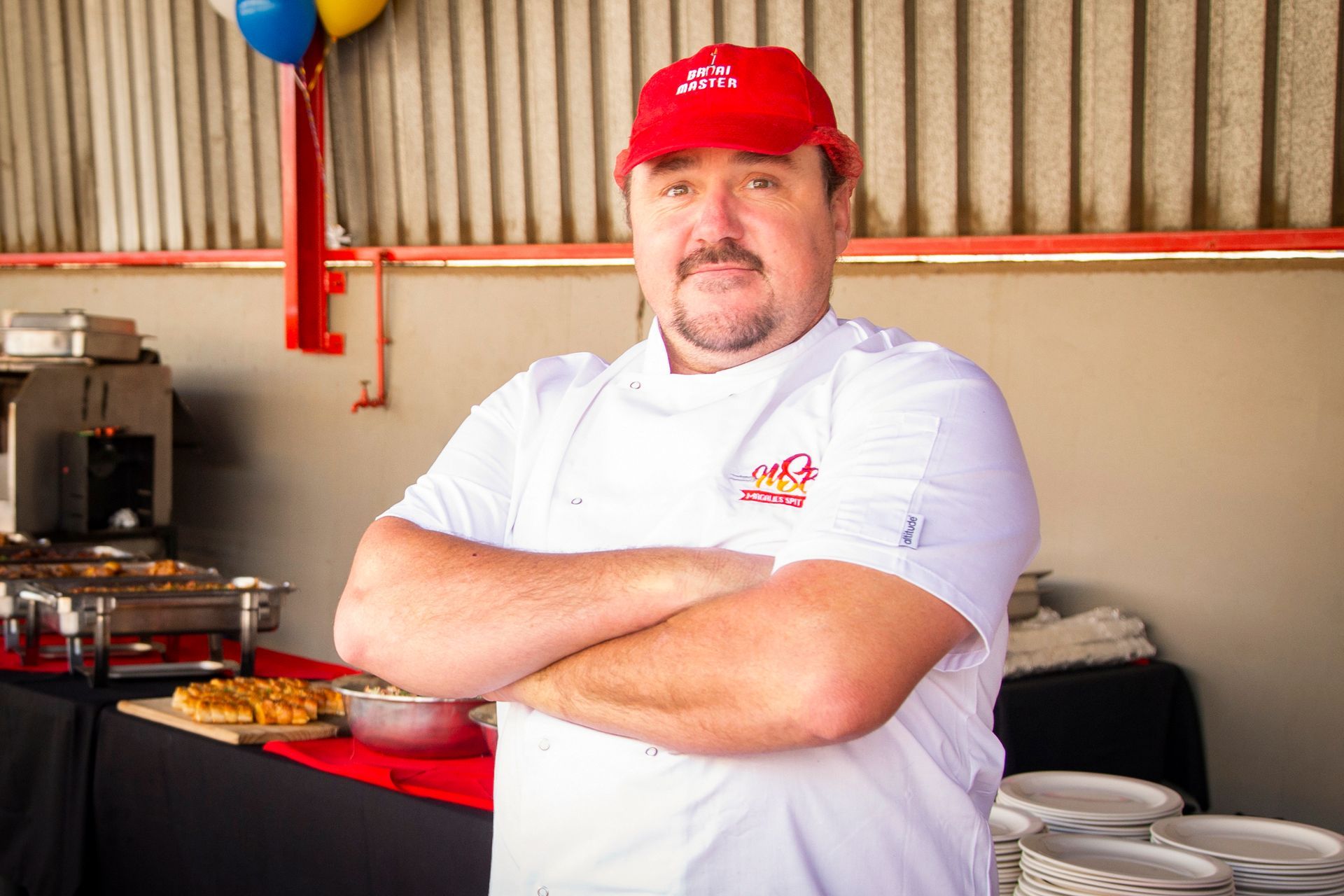 Chef with arms crossed, wearing a red hat and white chef coat. Standing in front of food on a table.