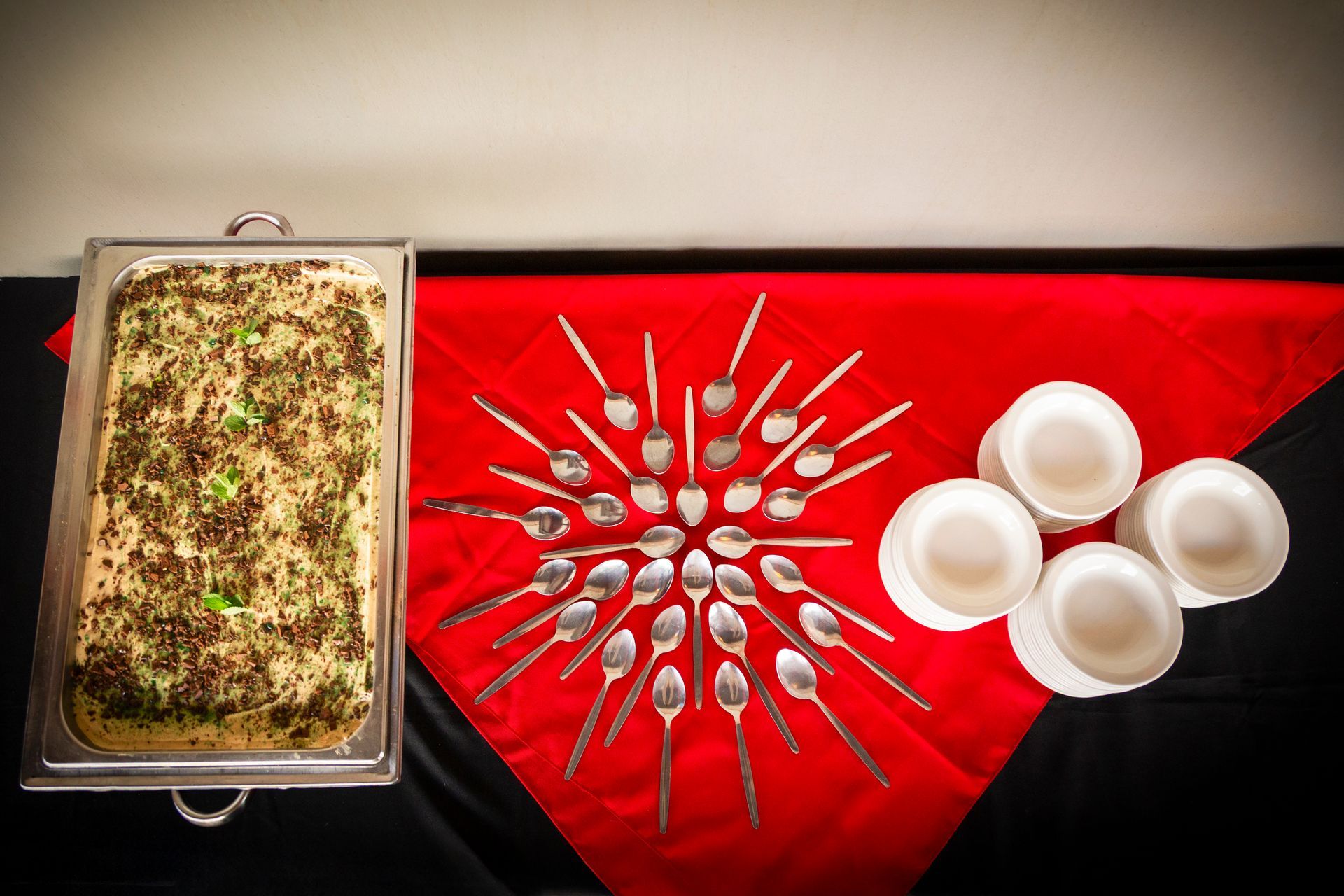 Catered food display: casserole dish, spoons arranged like sunburst, white bowls on red and black tablecloth.