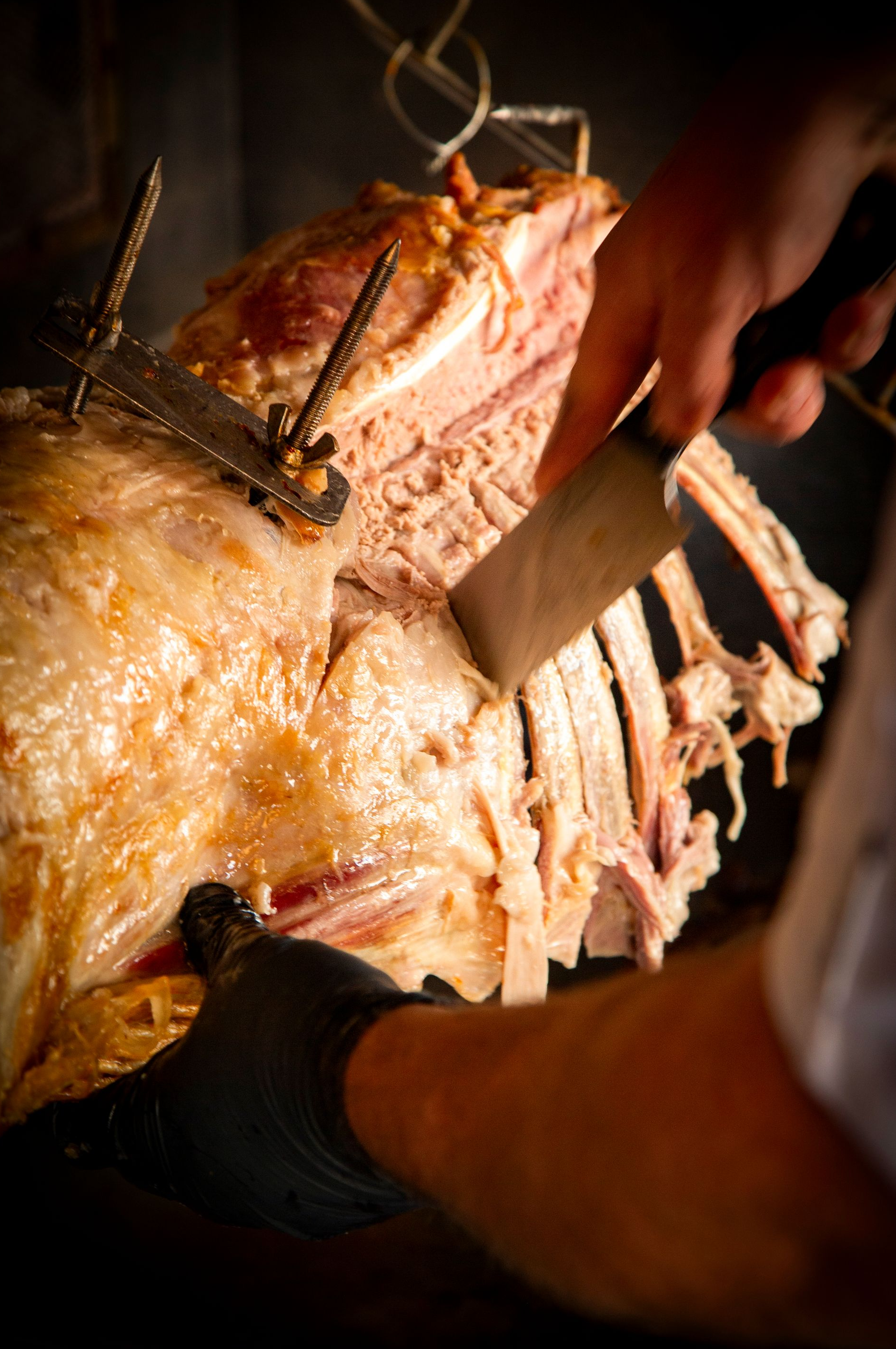Butcher carving roasted meat, ribs visible. Black-gloved hand with a knife, warm lighting.