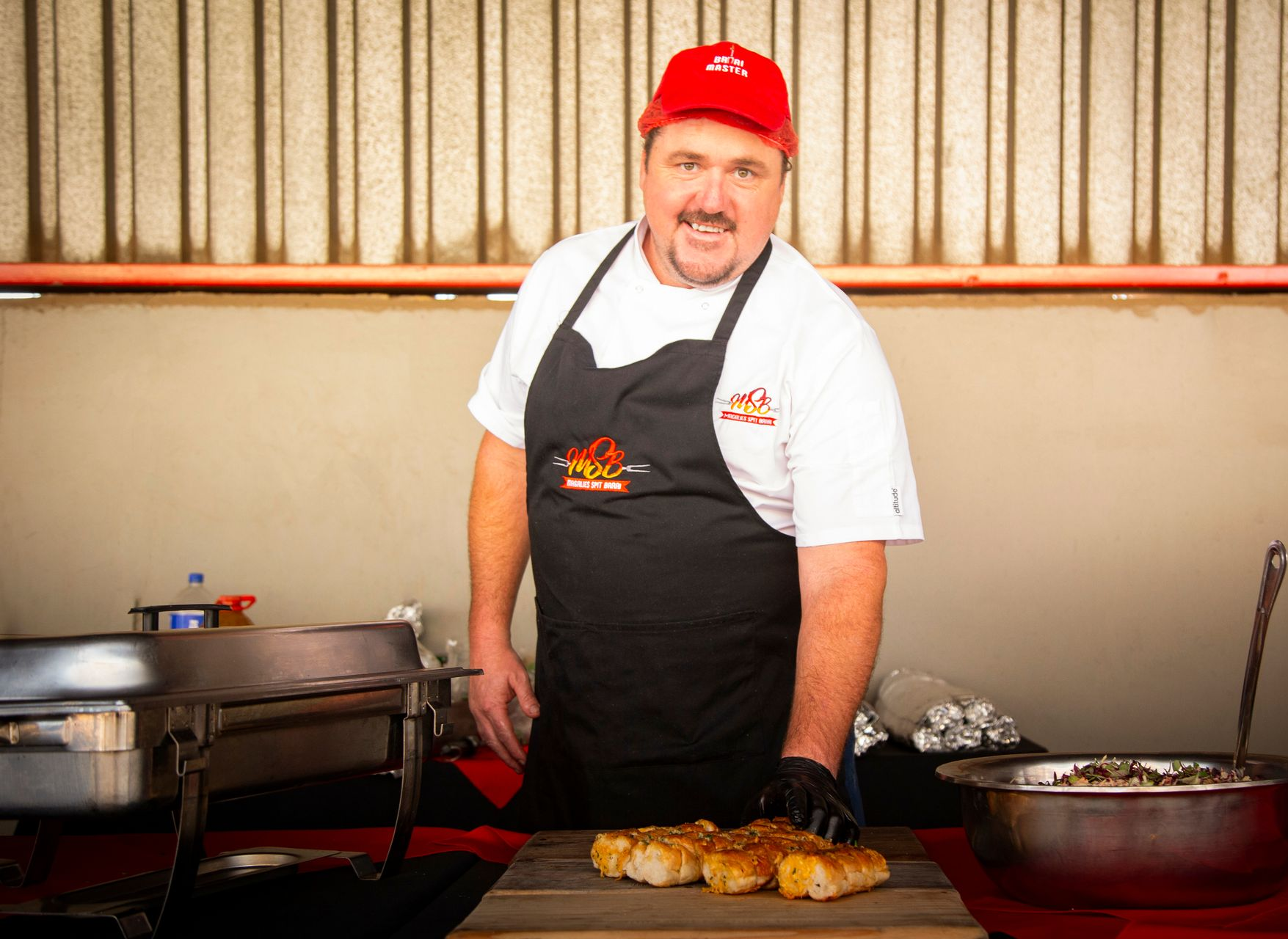 Man in red hat and apron stands at a food stall, smiling. He is holding food on a wooden board.