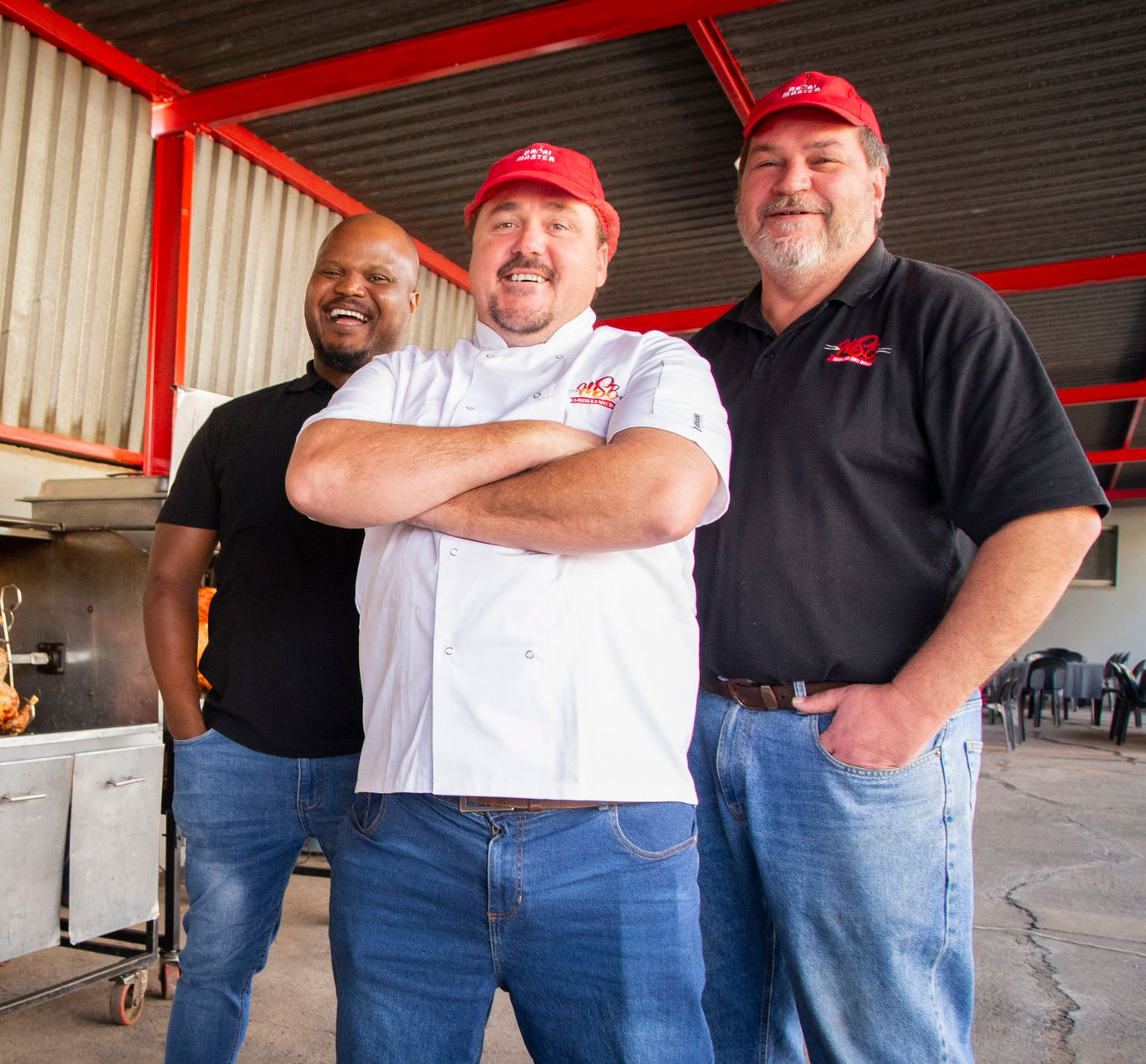 Three men in red hats smile; chef in white coat with crossed arms, others in casual shirts and jeans, near a rotisserie.