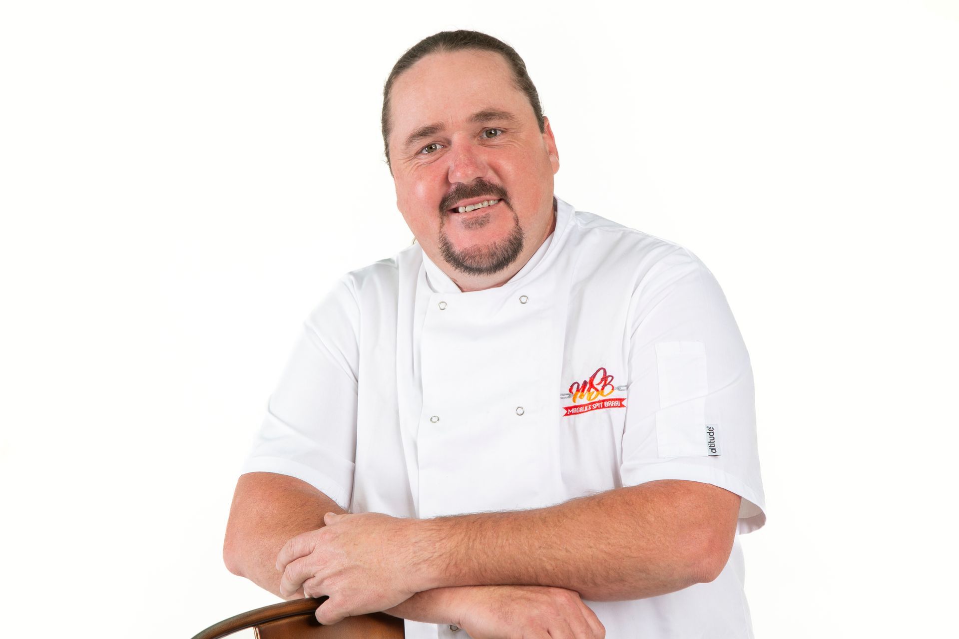 Chef in white coat smiling, leaning on a wooden object.