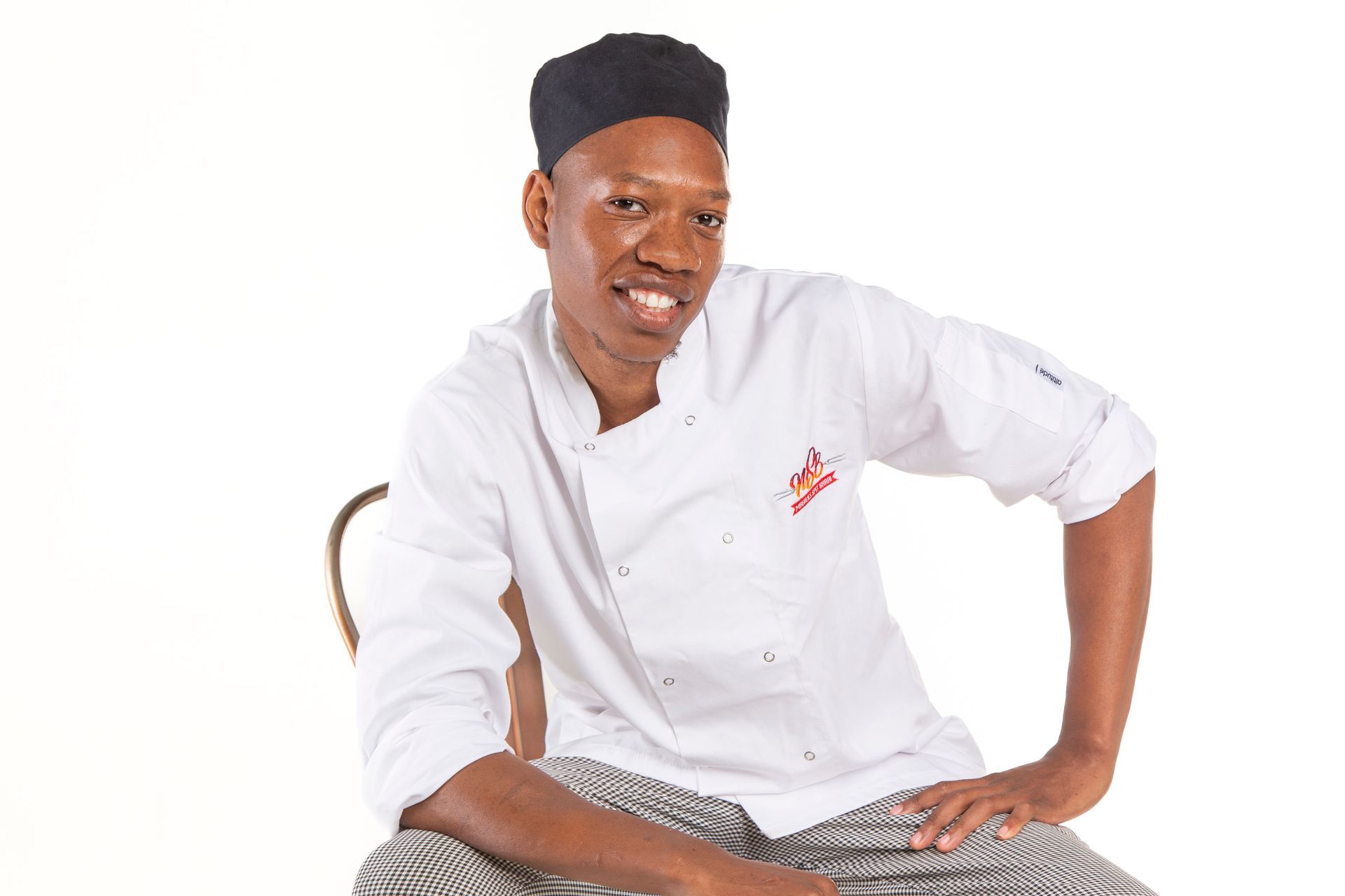 Young Black chef in white coat and black hat smiles, sitting on chair.
