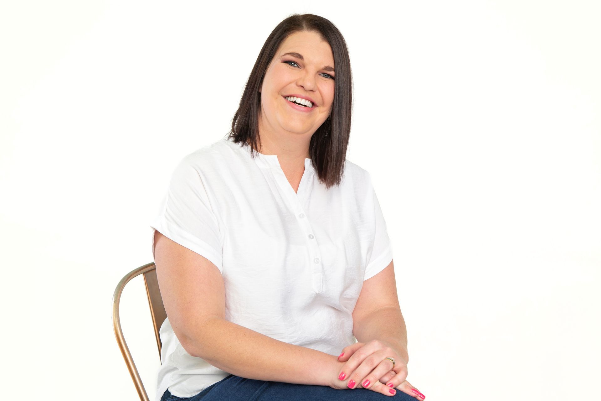 Woman sitting, smiling, wearing a white shirt and blue jeans, hands clasped in lap, against a white background.