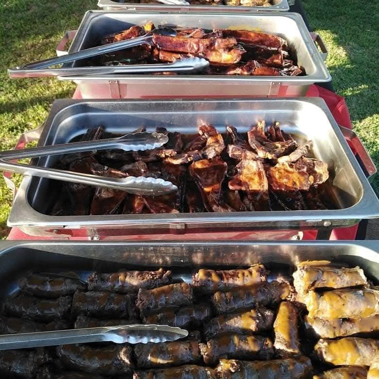 BBQ food: ribs, chops, and sausages in metal trays with tongs on a grassy area.