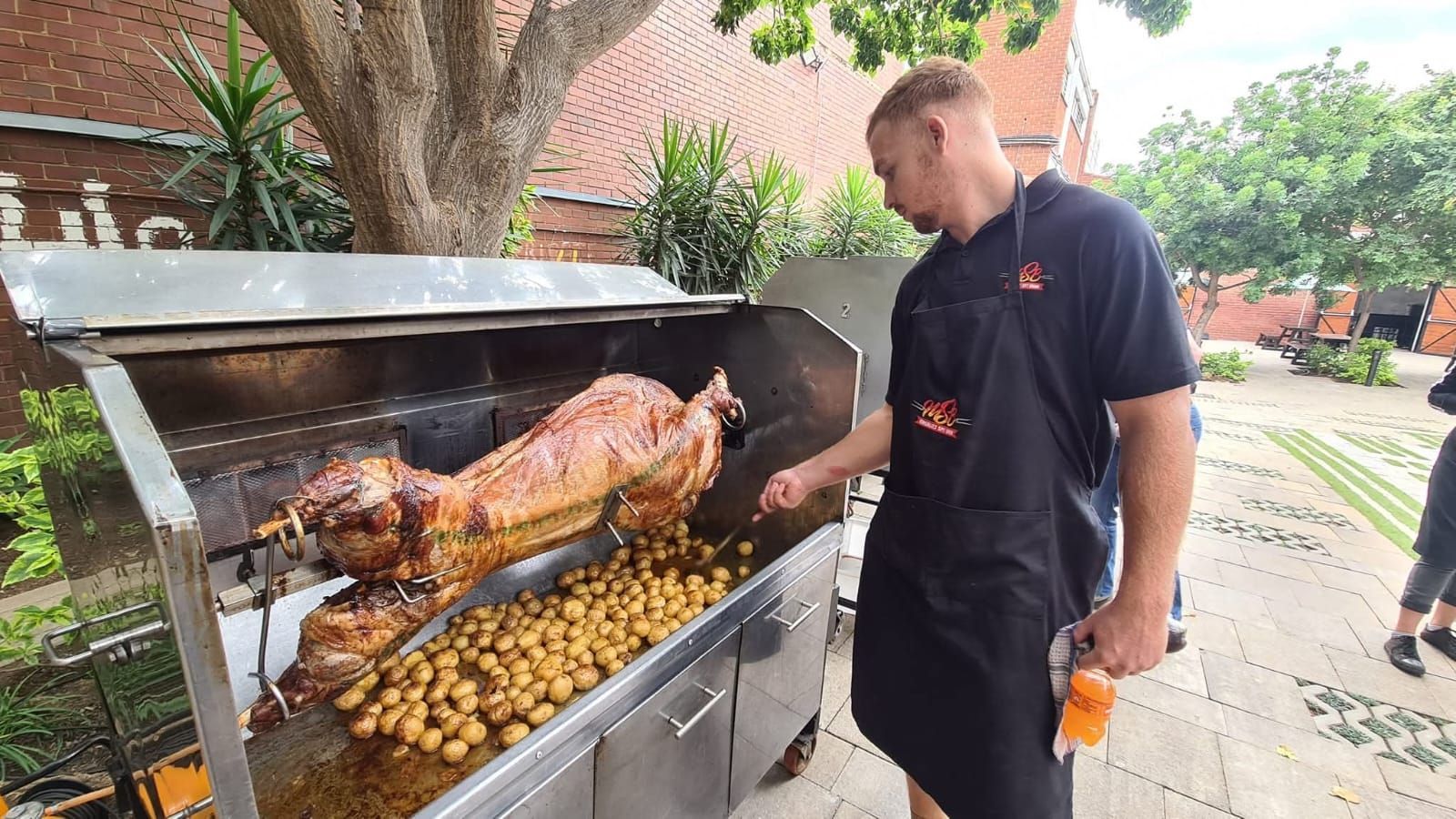 Man tending a roasted pig with potatoes in an outdoor rotisserie oven.