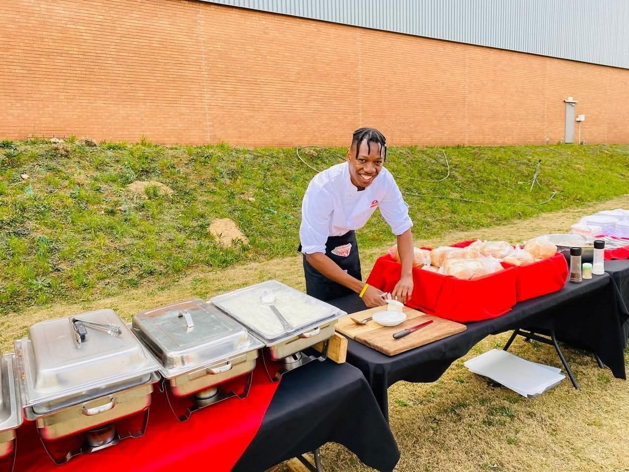 Chef smiling, prepping food at an outdoor catering table with chafing dishes and ingredients.